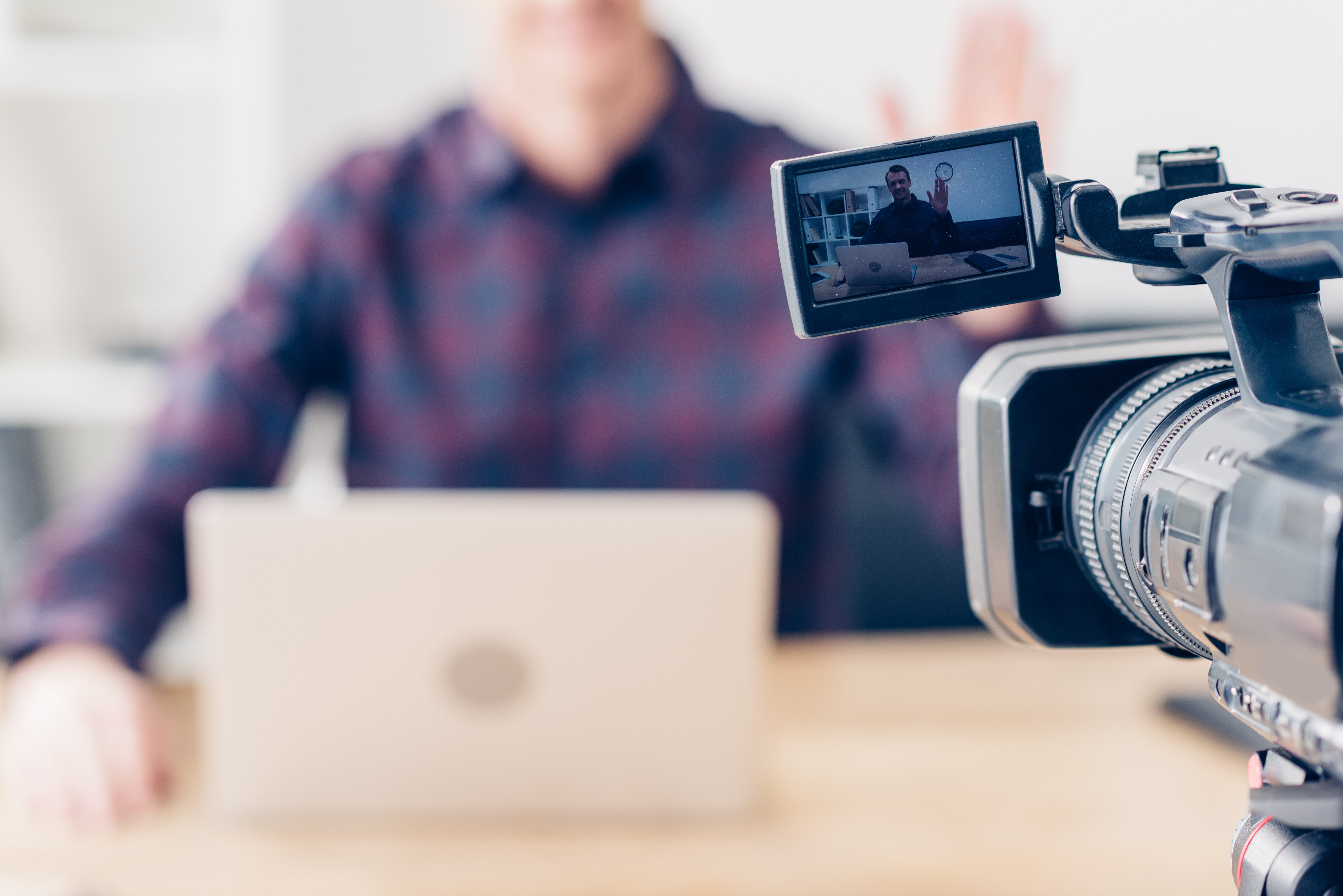 Person recording a video blog, with the cameras screen showing the individual waving while seated in front of a laptop.