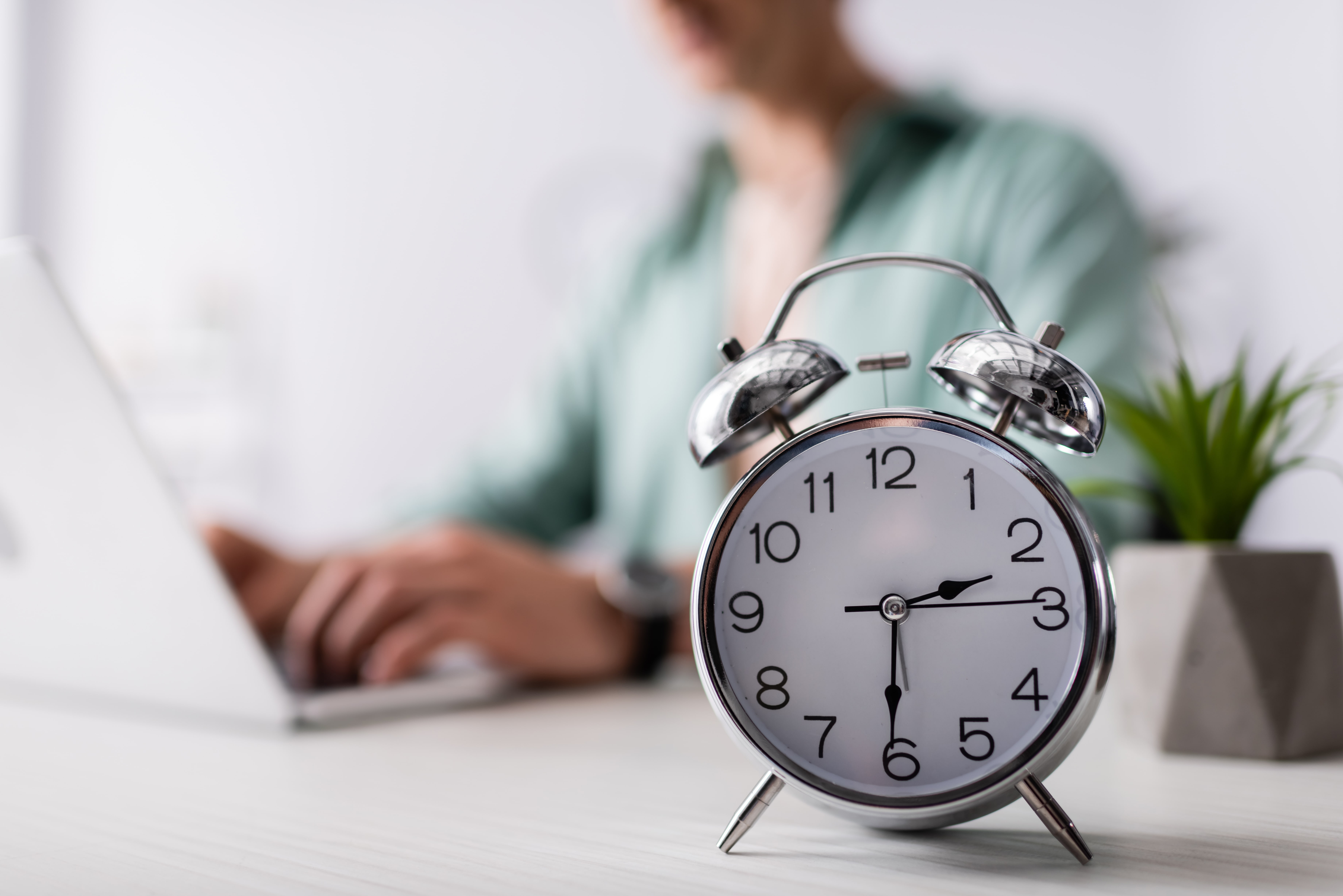 Selective focus on an alarm clock on a desk, with a man typing on a laptop in the background.