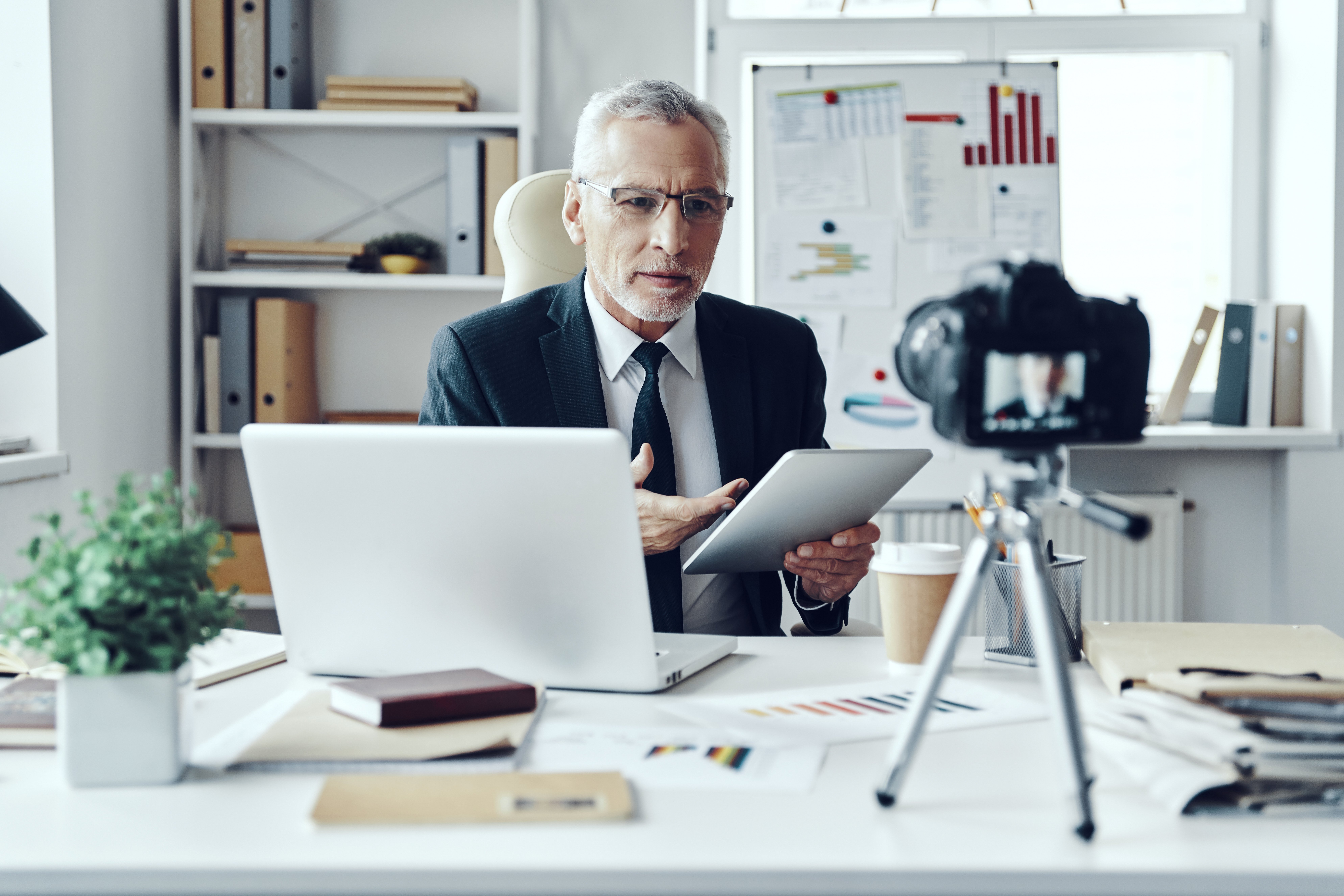 Senior man in a business suit using a tablet, sitting at a desk with a laptop, being recorded on a camera.