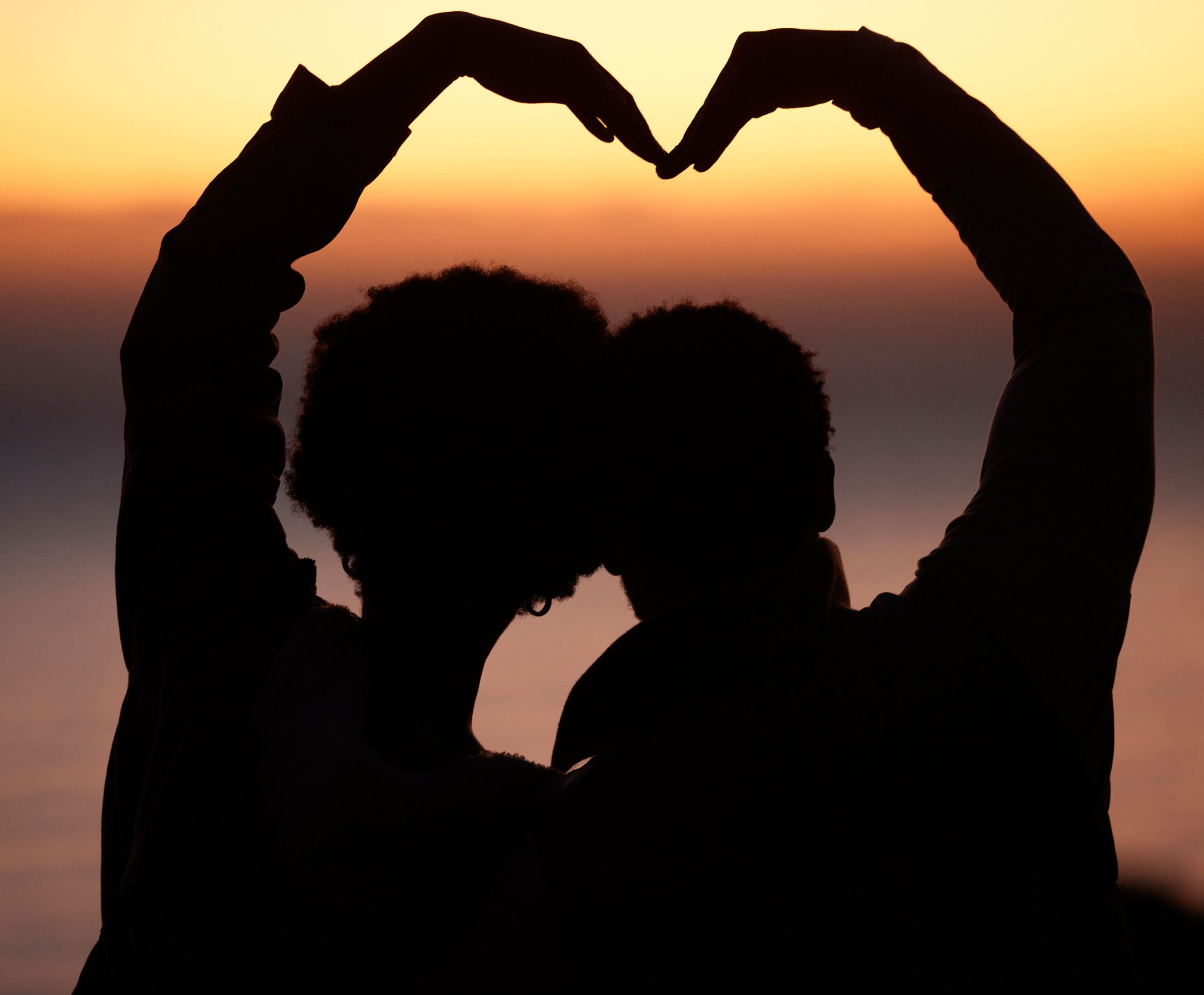 Silhouette of a couple forming a heart shape with their hands against a sunset background.-min