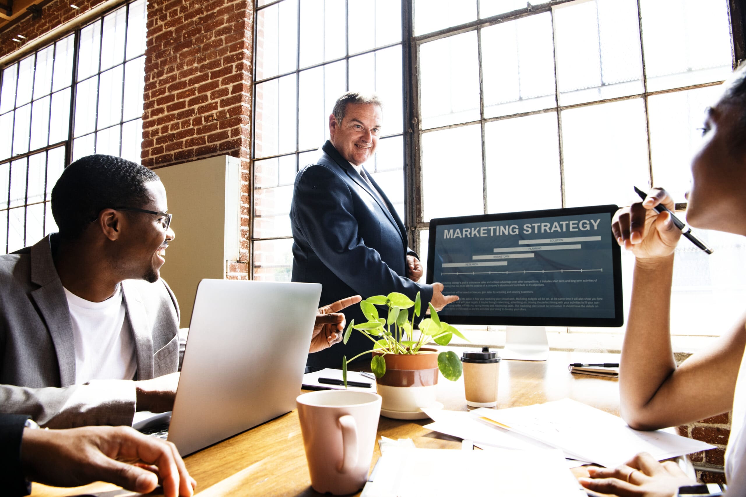 Smiling businessman presents marketing strategy on a screen to diverse colleagues in a sunlit, brick-walled office.-min