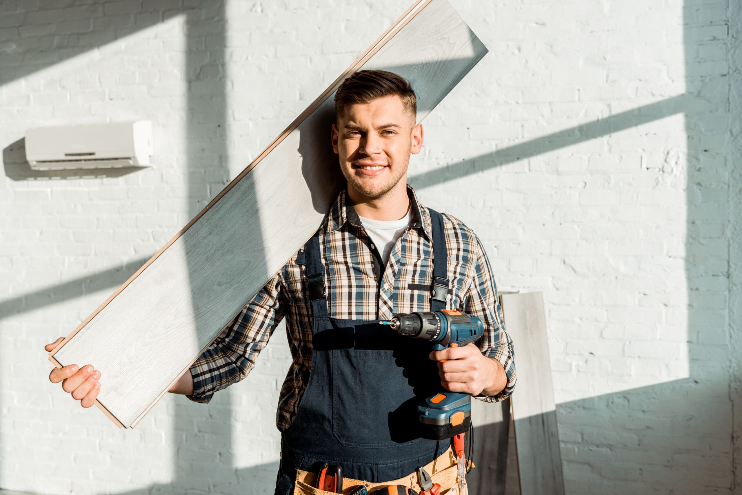 Smiling man in a plaid shirt holding a wooden board and a drill, ready for installation-min