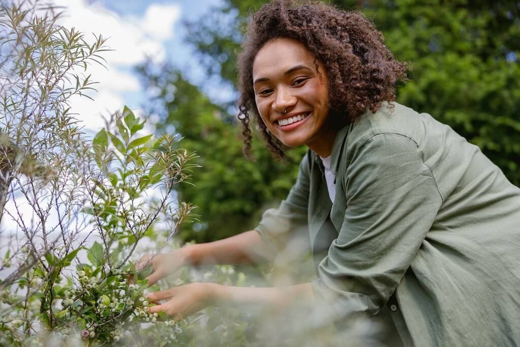 Smiling woman in a green shirt tending to plants in a garden on a sunny day.-min