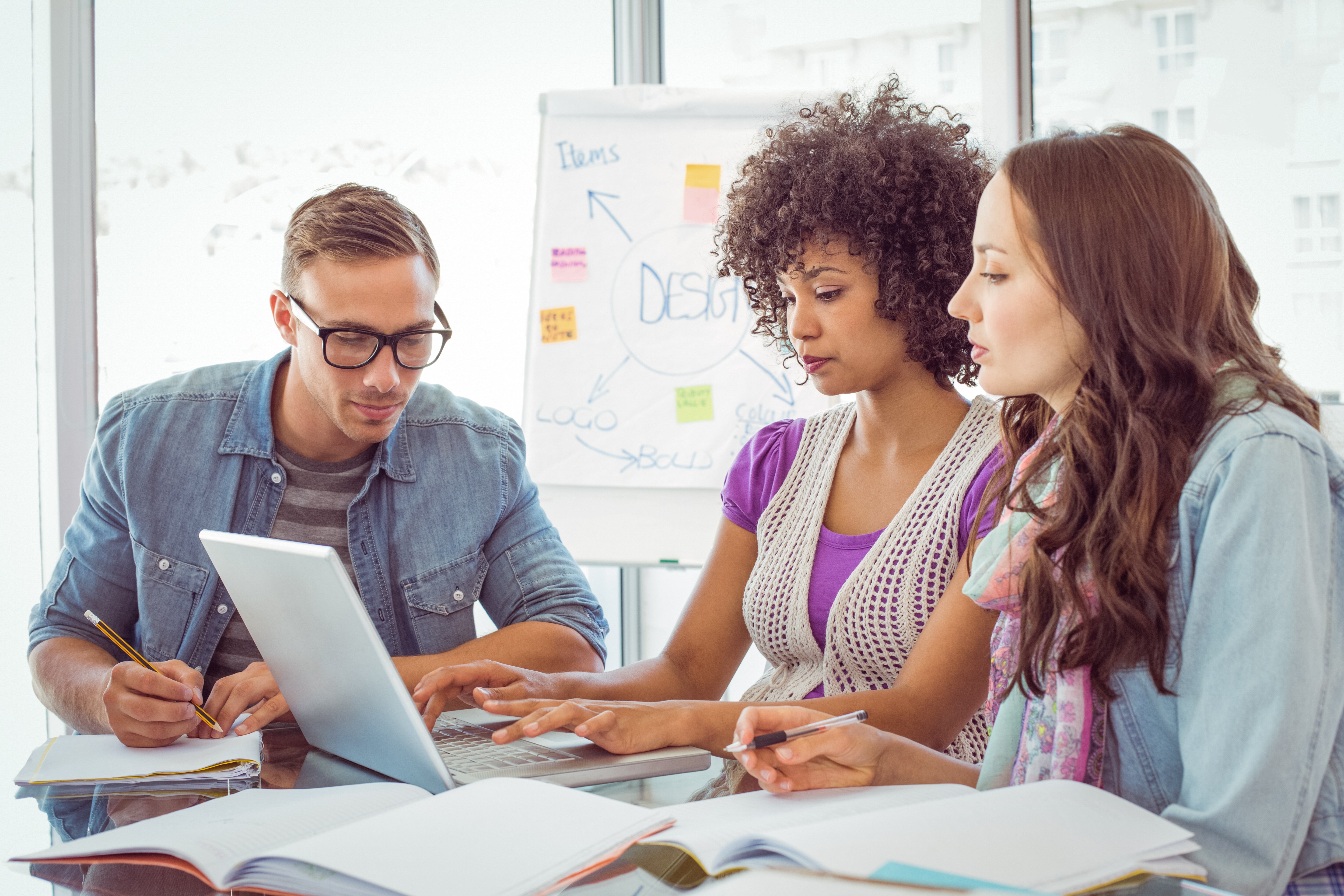 Three students collaborate at a table, using a laptop and taking notes, with a design brainstorming chart on a flipchart in the background.