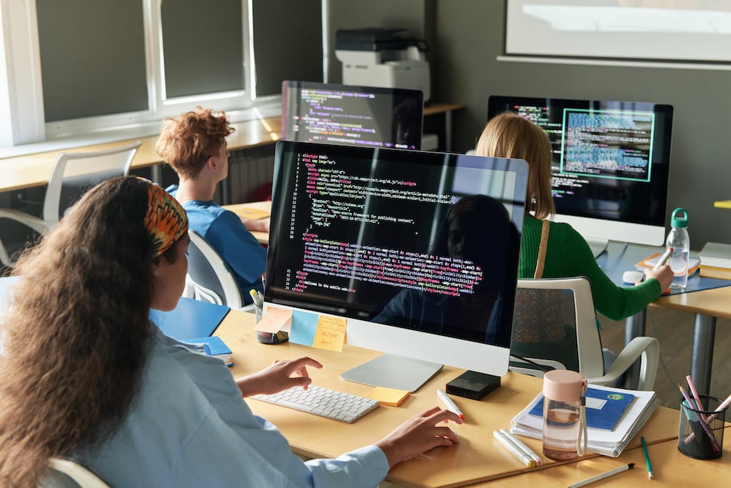 Three students focus on writing code during a programming lesson, each working at their computer stations with lines of code displayed on their screens.