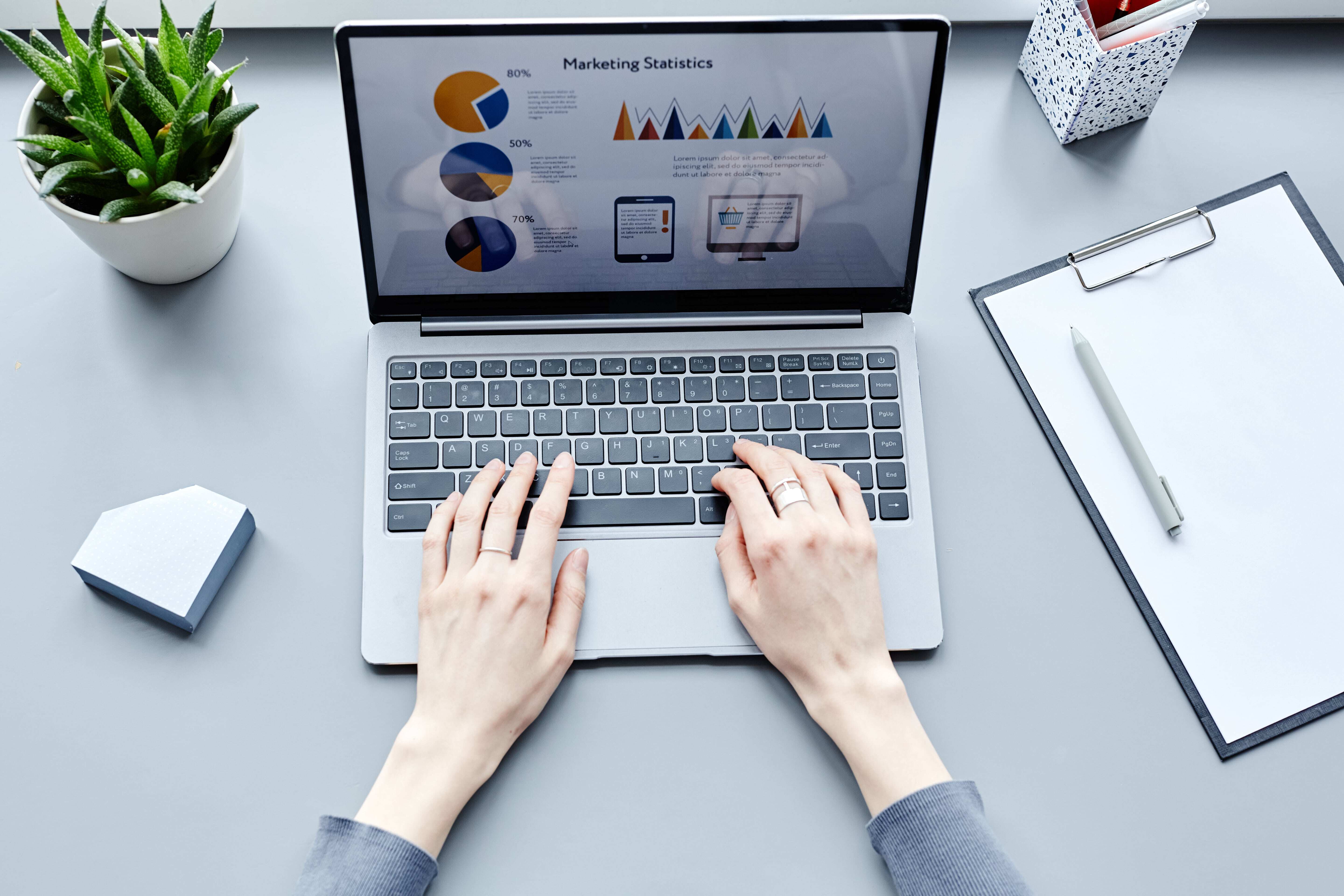 Top view of hands typing on a laptop with marketing statistics displayed on the screen, a clipboard, and a potted plant nearby