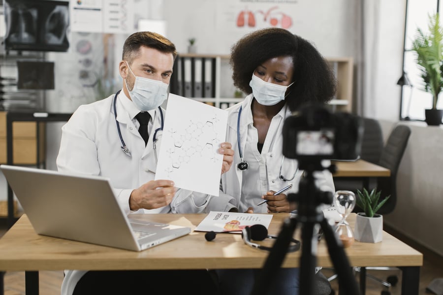 Two masked doctors discuss a research diagram while recording a video at a desk with a laptop and microphone, suggesting a medical presentation.