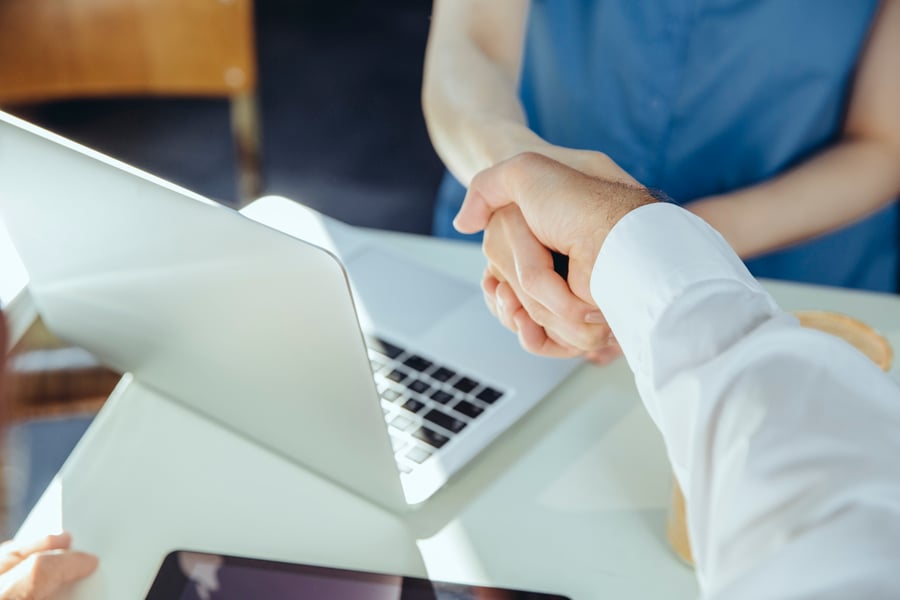 Two medical device marketers shake hands over a table with a laptop, symbolising a business agreement or partnership in a professional setting. 