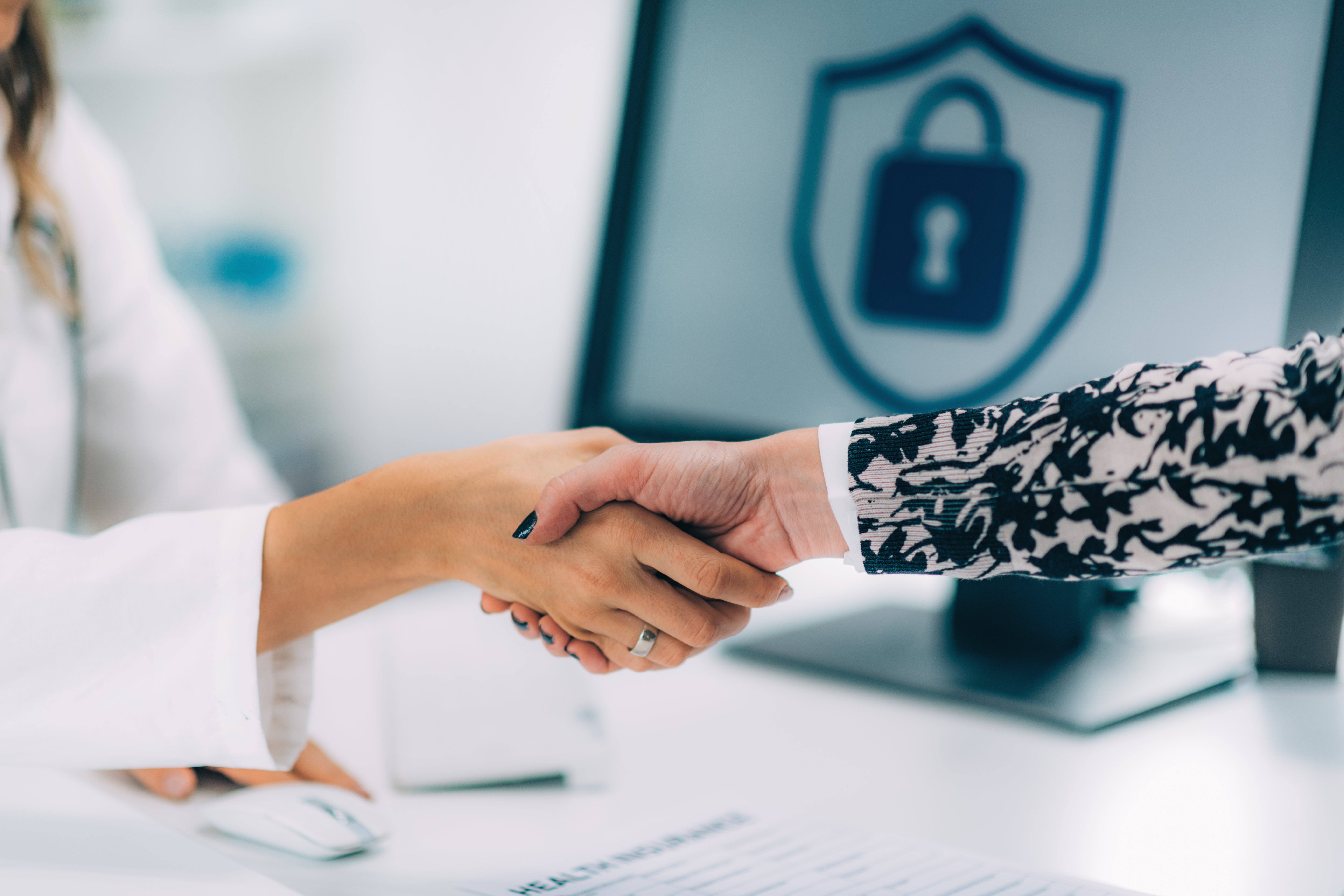 Two people shaking hands over a desk with a computer screen displaying a lock symbol in the background, symbolising data security.