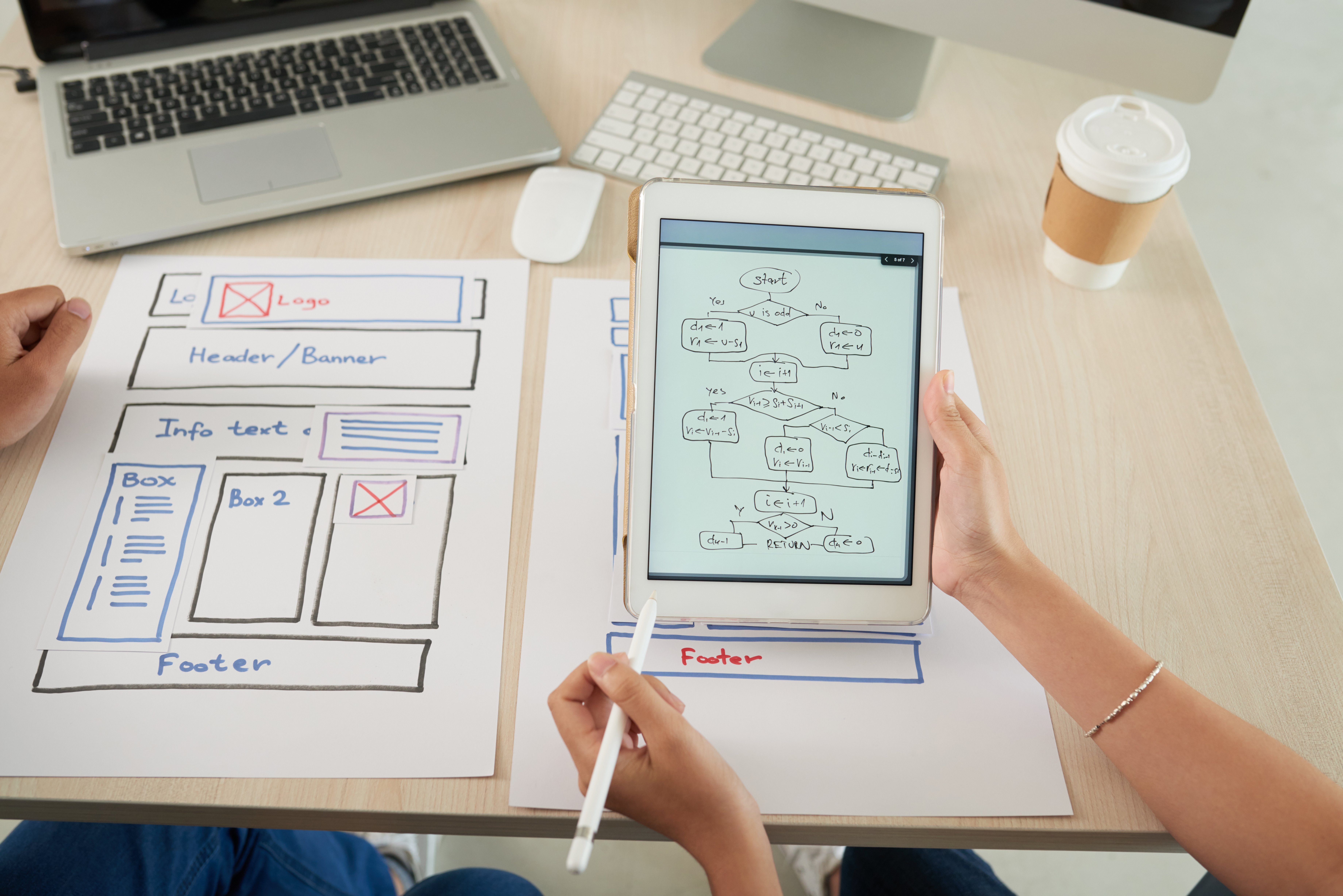 Two people work on a website layout, with one holding a tablet showing a flowchart and the other reviewing a wireframe sketch on paper.