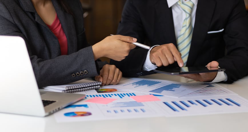 Two professionals in suits discuss data while pointing at a tablet, with charts, graphs, and a laptop on the table, indicating a business meeting.