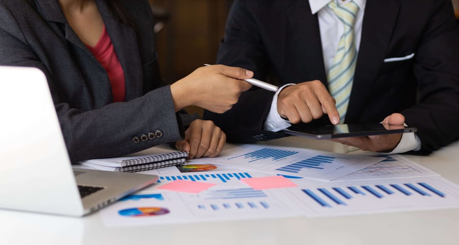 Two marketers in suits discuss data while pointing at a tablet, with charts, graphs, and a laptop on the table, indicating a business meeting.
