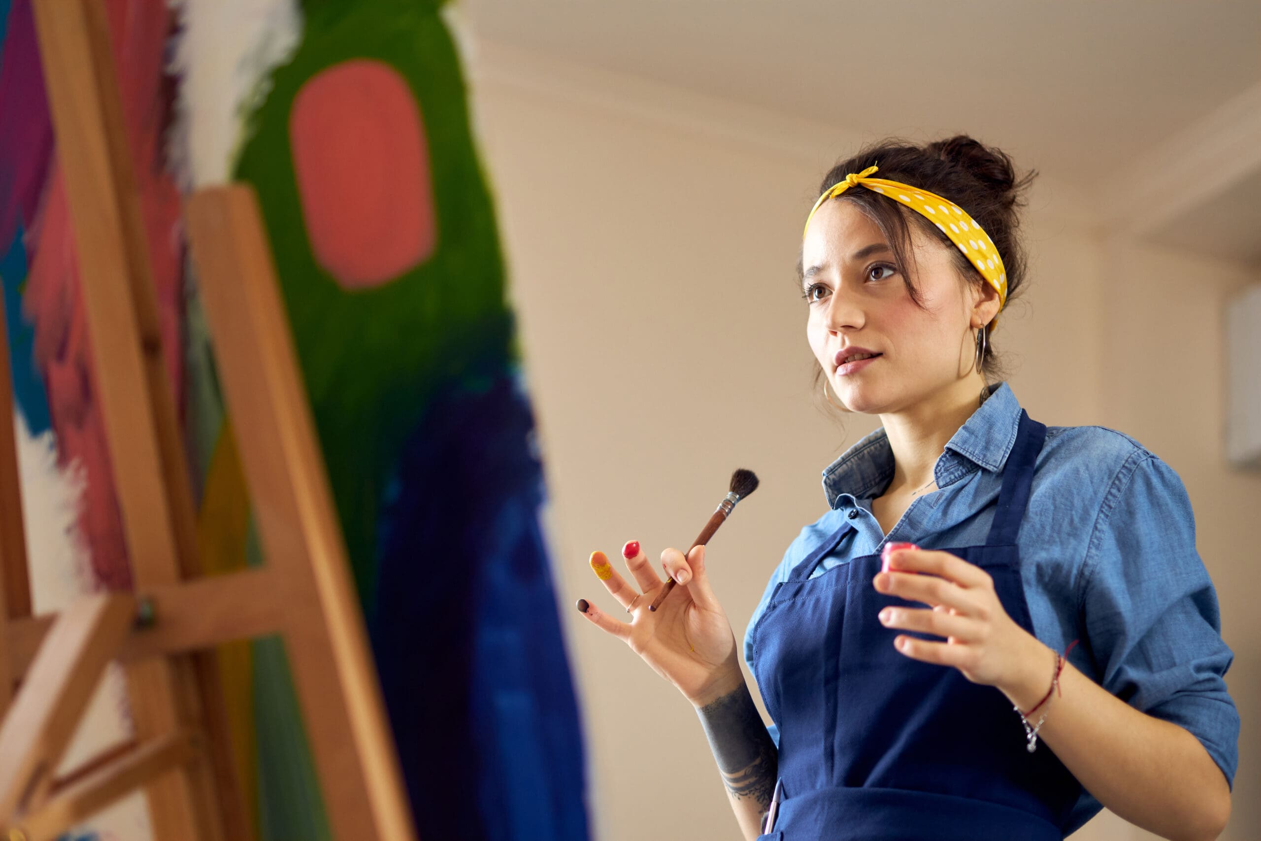 Young woman in a denim shirt and apron holding a paintbrush, standing in front of a colourful canvas-min