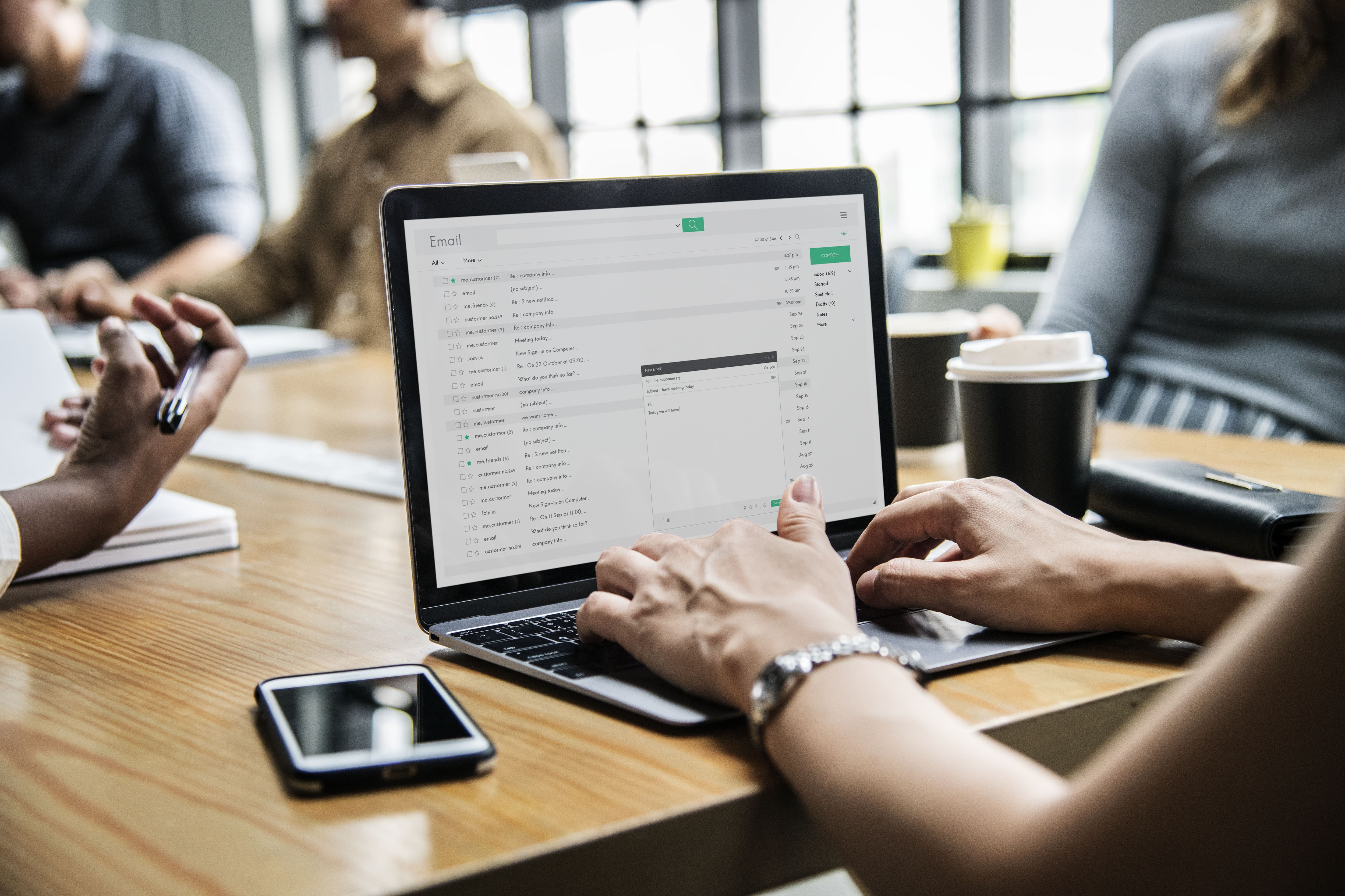 Person working on a laptop in a meeting, composing an email for a medical device marketing campaign, with colleagues discussing strategies in the background. 