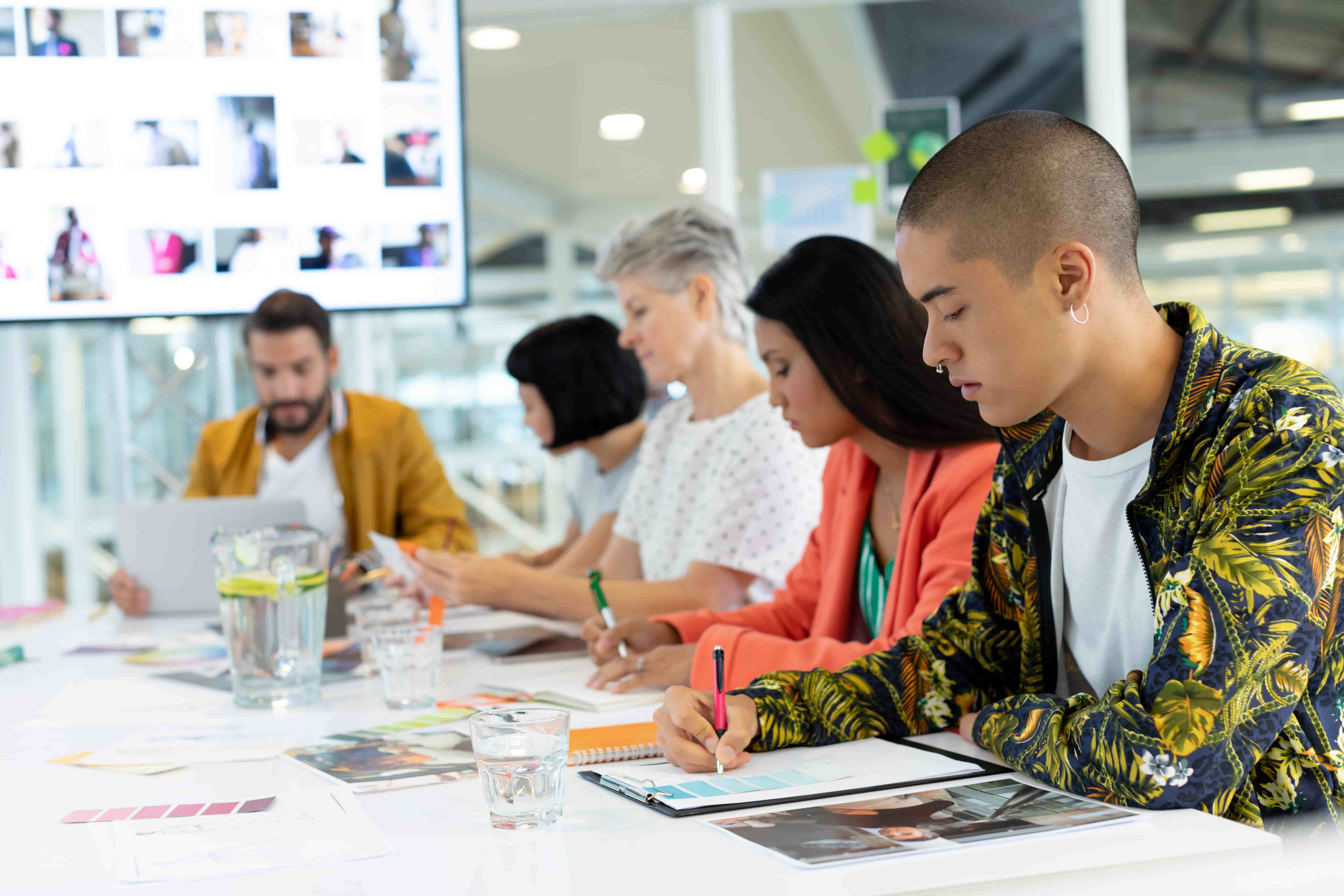 A diverse group of marketers sits at a table, reviewing documents and working on laptops in a bright, modern office setting.