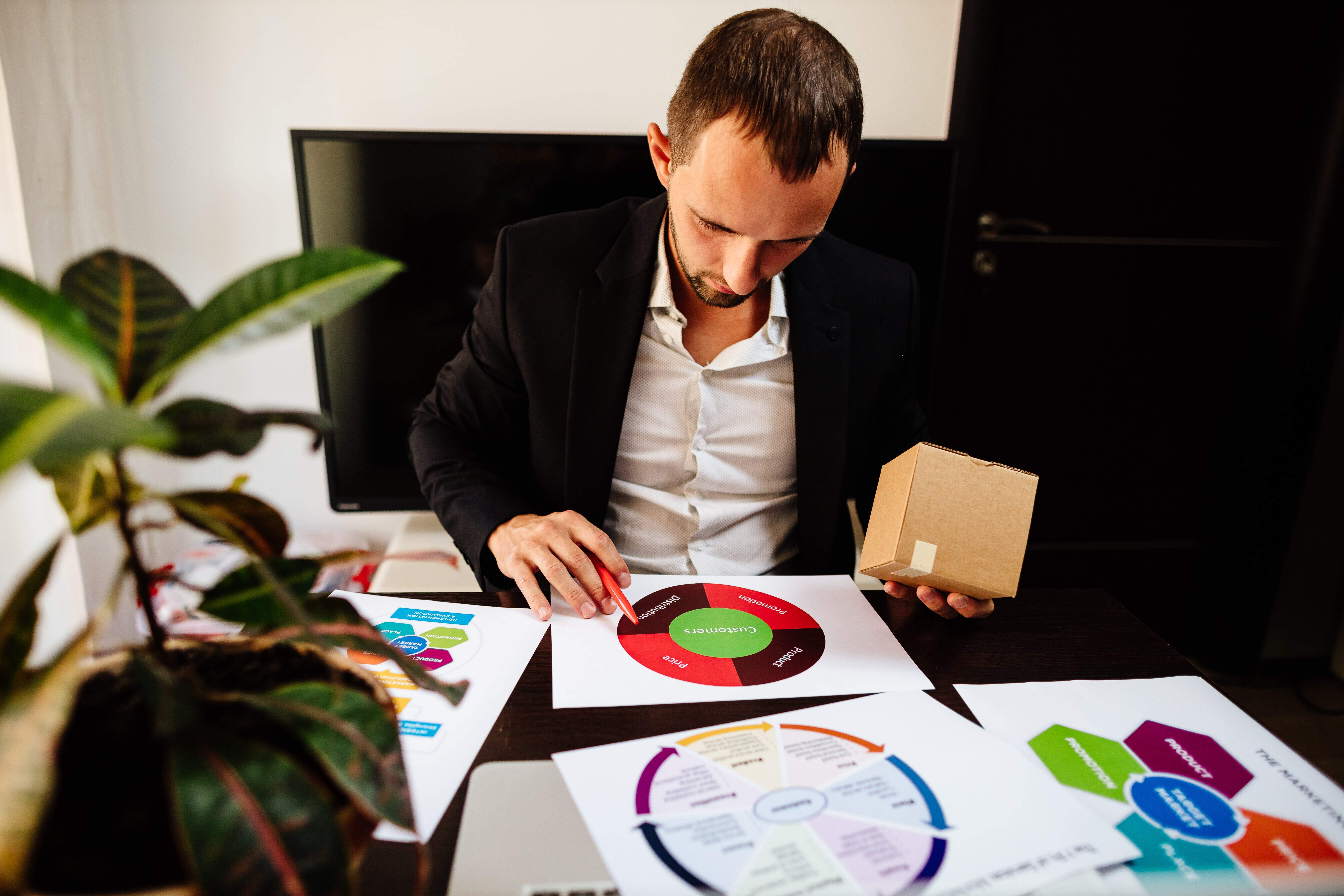 A man in a suit studies a circular marketing strategy diagram at a desk, holding a small cardboard box. 