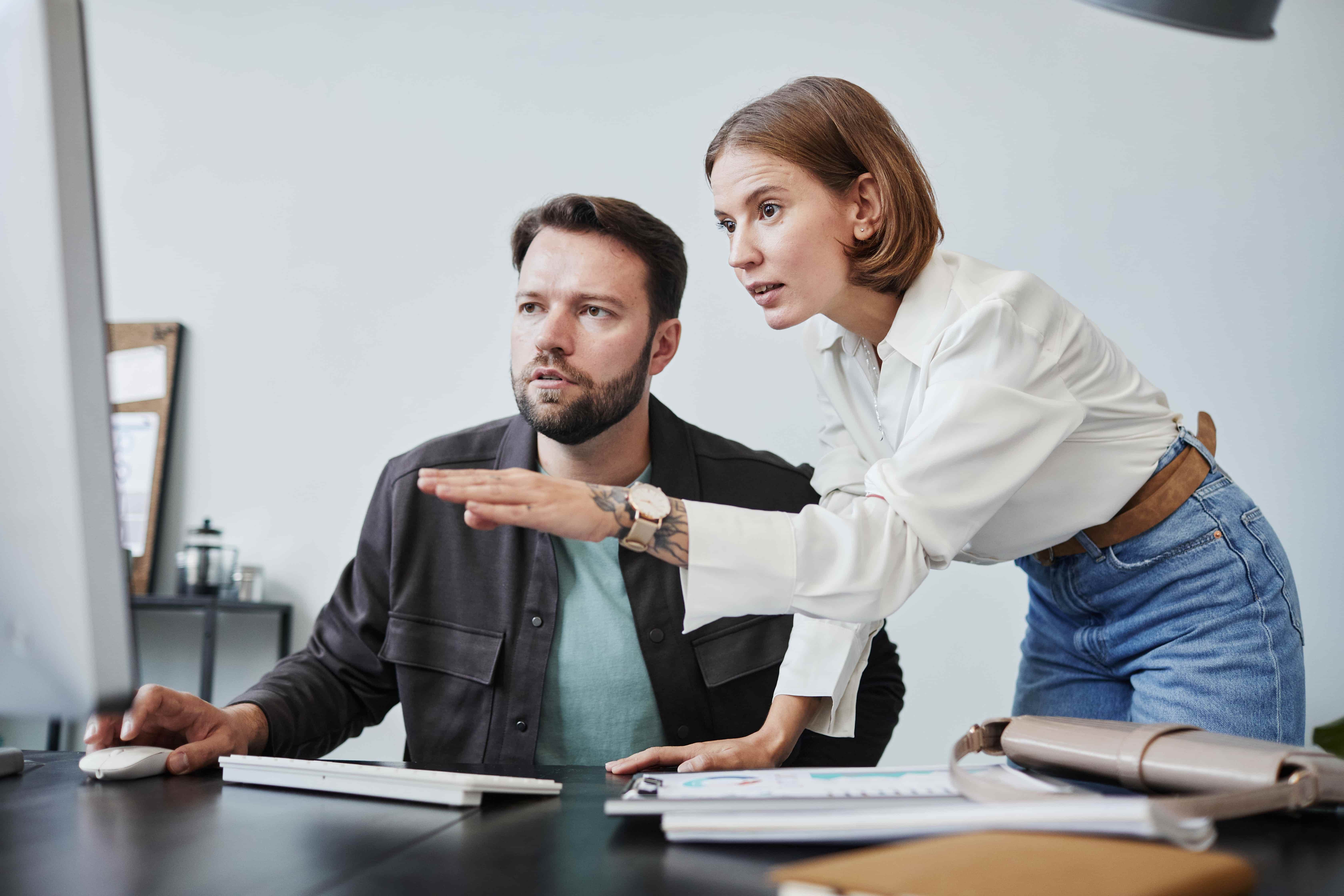 Two colleagues collaborate at a desk, with one pointing at images on a computer screen while the other observes closely. 