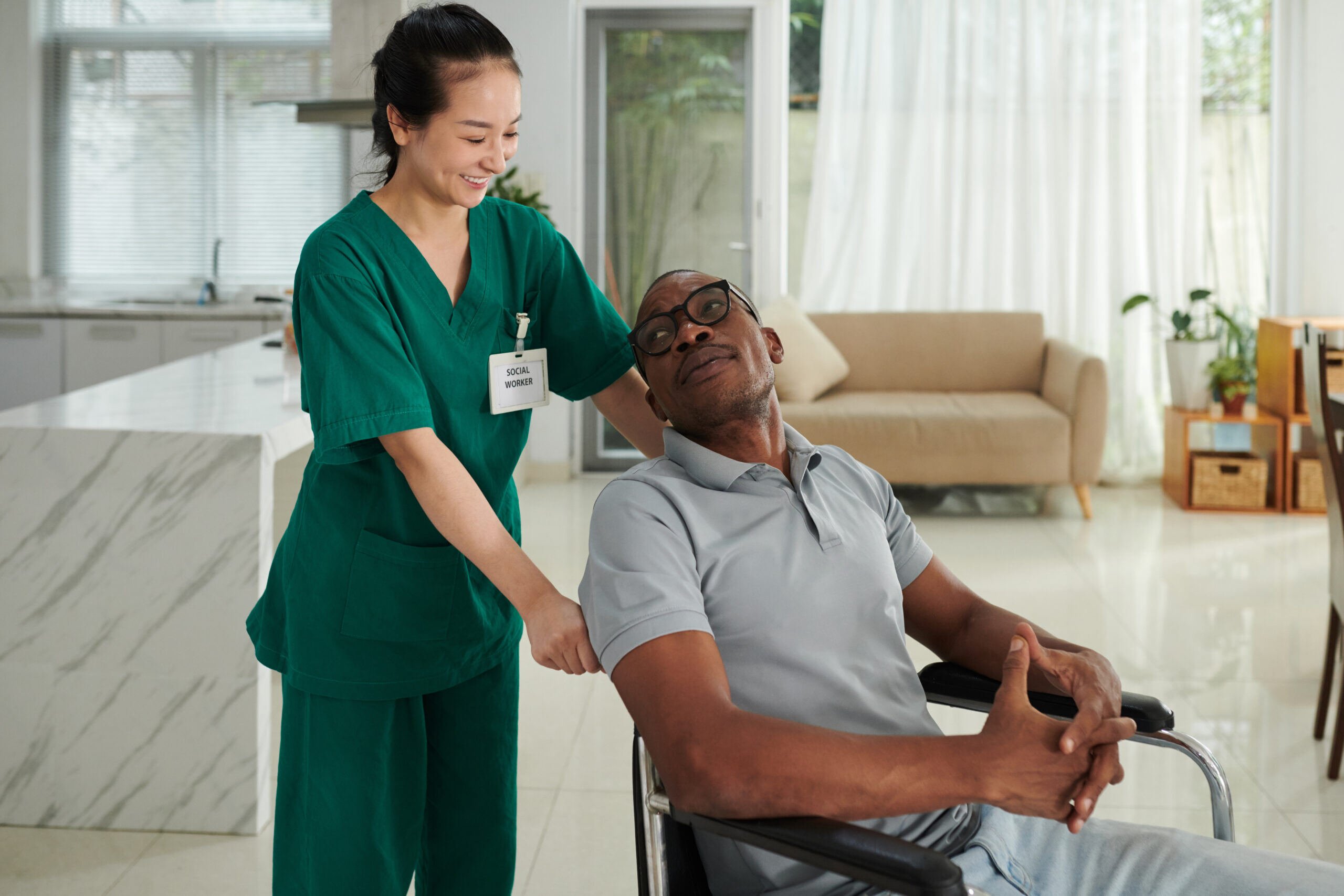 Female social worker in green scrubs smiling while pushing a male patient in a wheelchair.