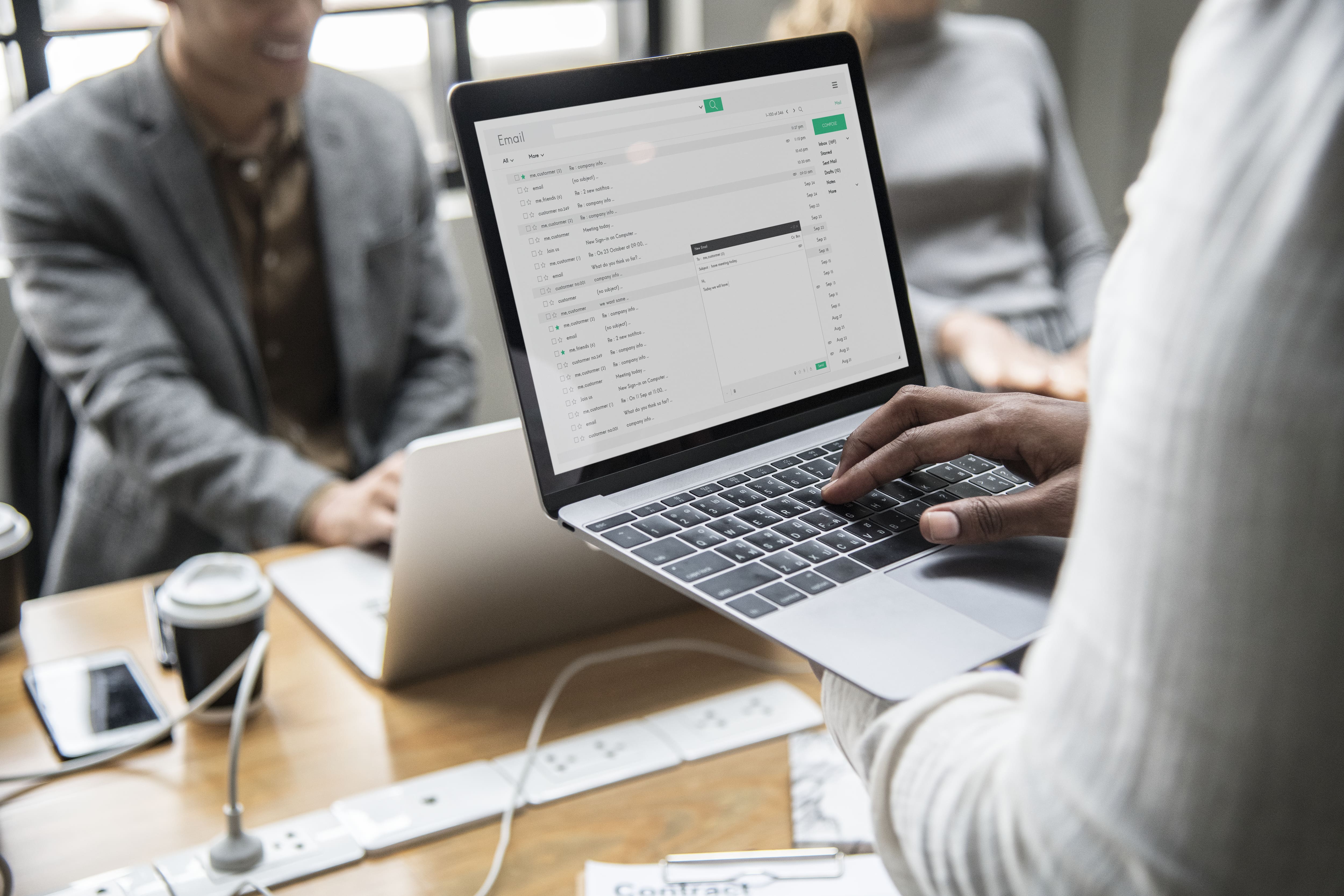 Person holding a laptop showing an email inbox screen, with two colleagues in the background. The scene is set in a modern office. 
