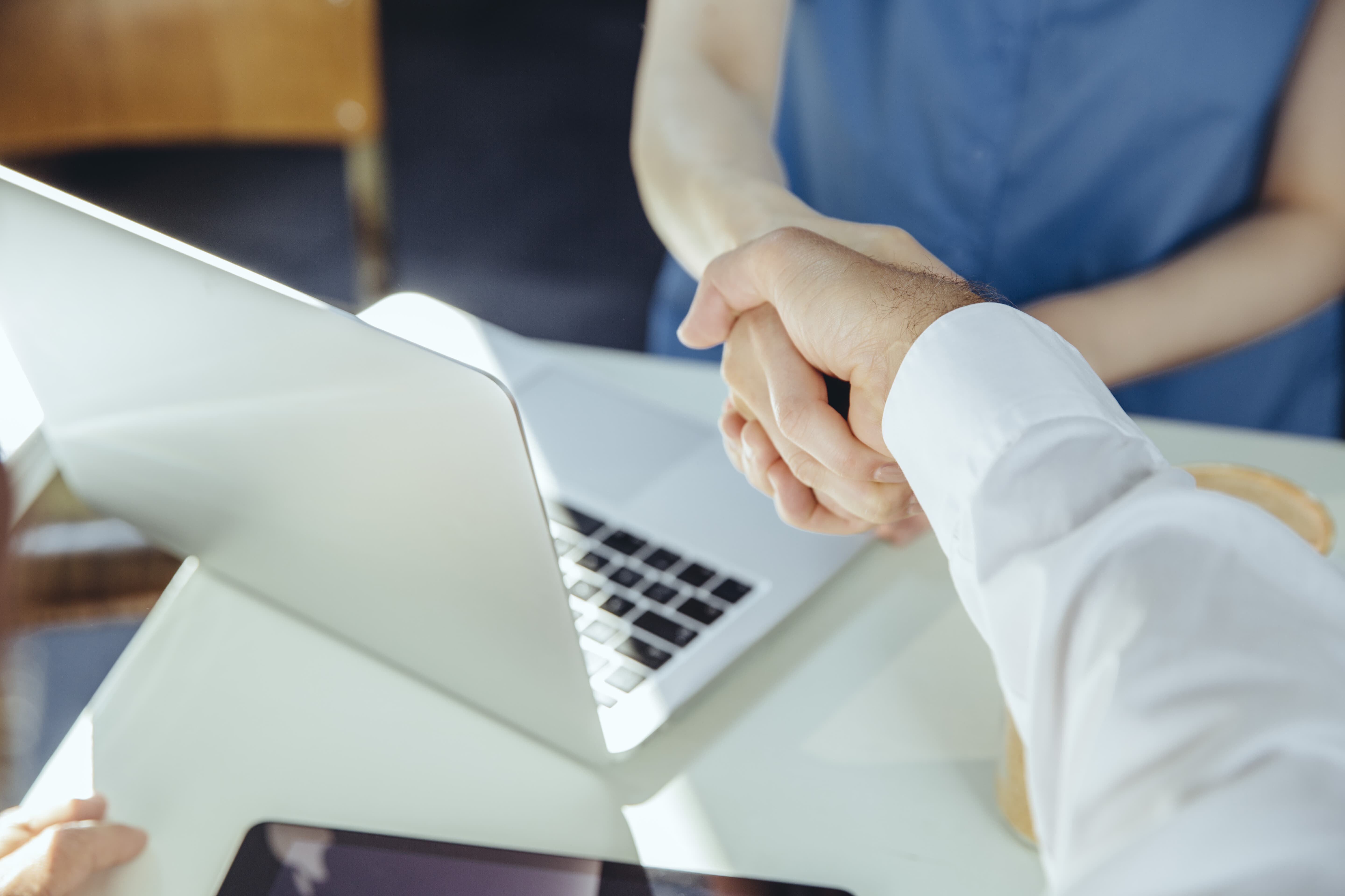 Two medical device marketers shake hands over a table with a laptop, symbolising a business agreement or partnership in a professional setting. 