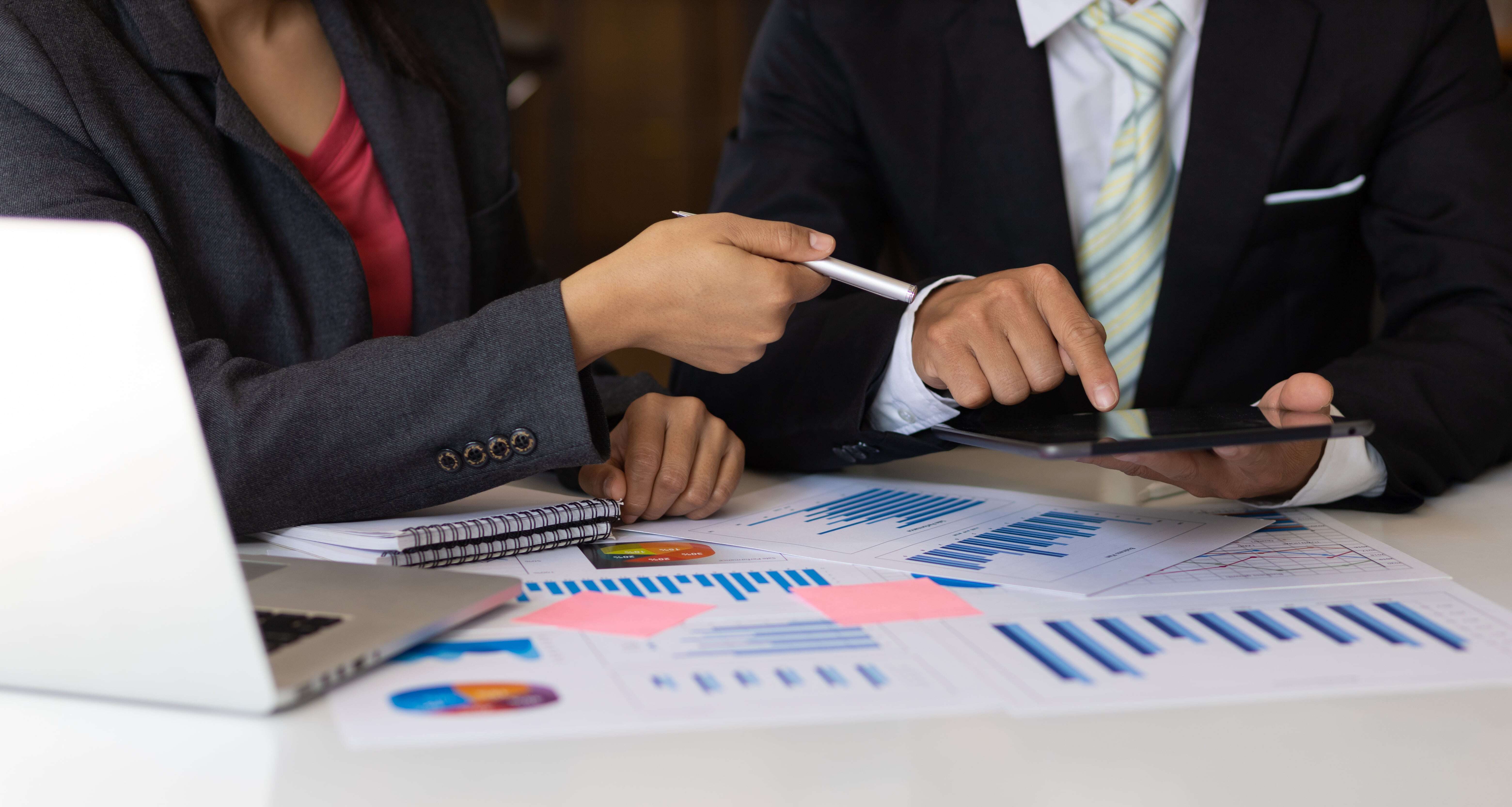 Two marketers in suits discuss data while pointing at a tablet, with charts, graphs, and a laptop on the table, indicating a business meeting.
